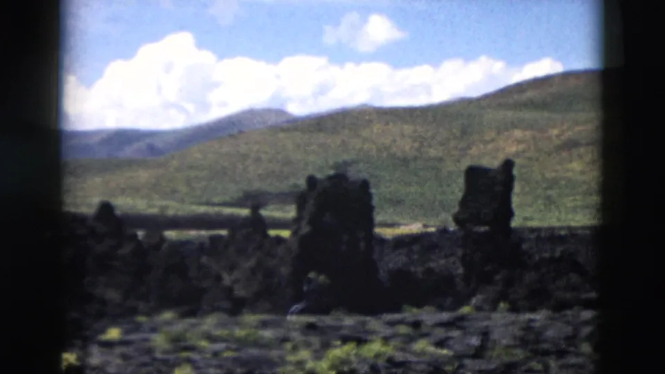 1958 Yellowstone frame showing a wider volcanic landscape with dark rocks, rolling green hills, and bright clouds.
