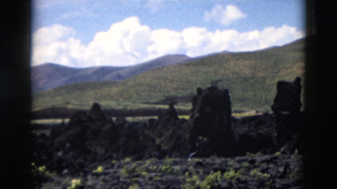 1958 Yellowstone 8mm frame showing black volcanic rock, sparse plants, distant hills, and a blue sky with clouds.