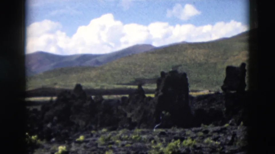 1958 Yellowstone 8mm frame showing black volcanic rock, sparse plants, distant hills, and a blue sky with clouds.