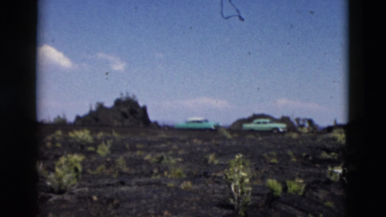 1958 Yellowstone frame with two turquoise cars parked on dark volcanic-looking ground beside sparse shrubs under a bright sky.