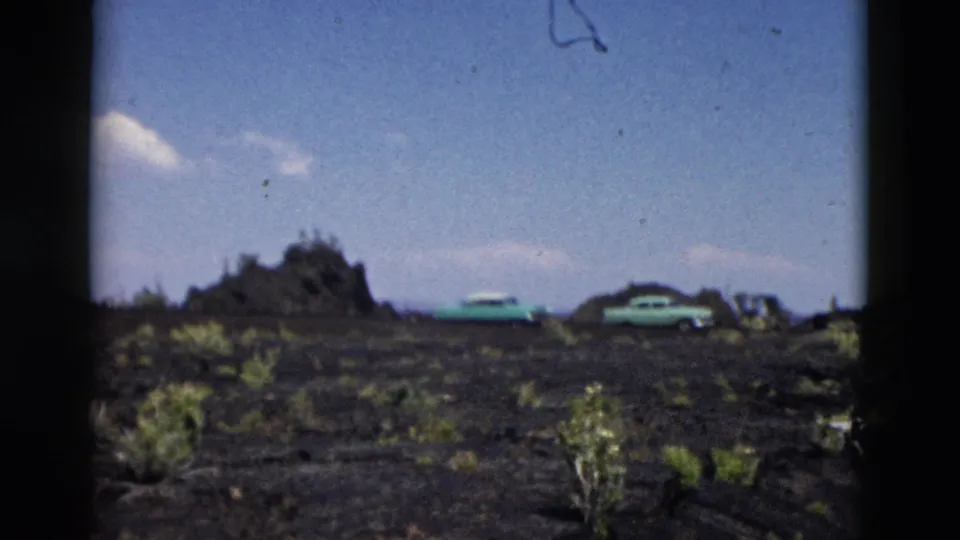 1958 Yellowstone frame with two turquoise cars parked on dark volcanic-looking ground beside sparse shrubs under a bright sky.