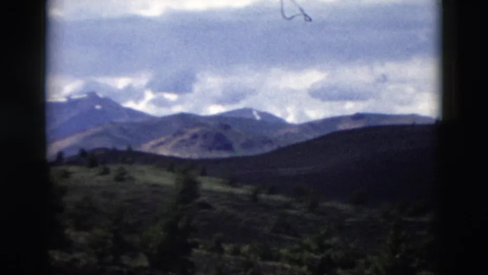 1958 Yellowstone frame showing a hazy mountain range under clouds, with dark rolling hills and sparse vegetation in the foreground.