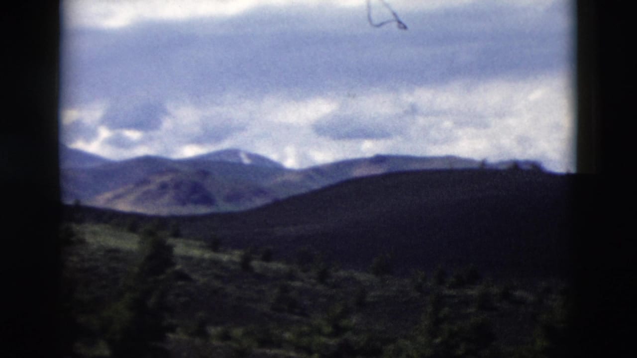 1958 Yellowstone home-movie frame showing distant mountains, shadowed hills, scattered evergreens, and a cloudy sky.
