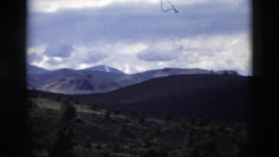 1958 Yellowstone home-movie frame showing distant mountains, shadowed hills, scattered evergreens, and a cloudy sky.
