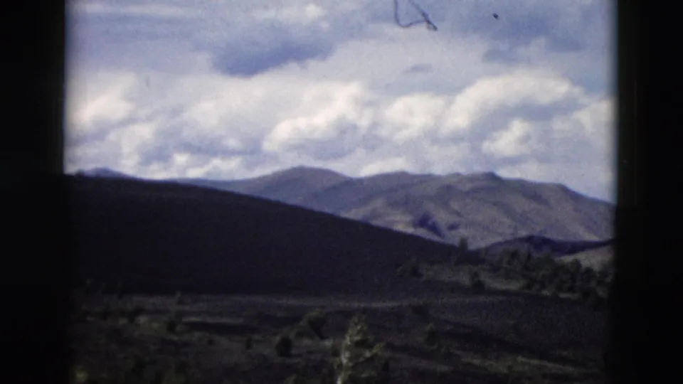 Yellowstone home-movie frame showing a mountain ridge, scattered evergreens, rugged ground, and soft white clouds.