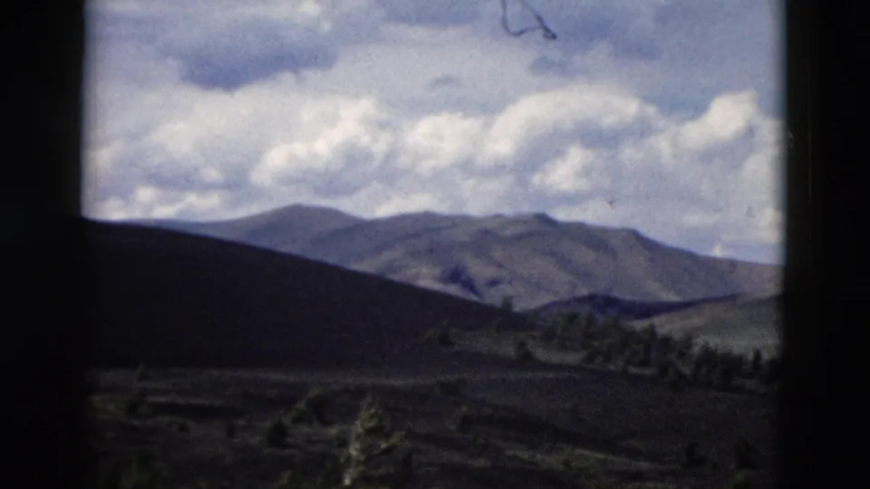 1958 Yellowstone-area 8mm frame showing layered mountain ridges, shadowed slopes, sparse trees, and cumulus clouds.