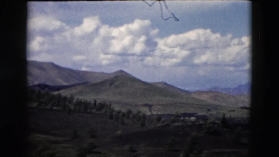 1958 Yellowstone-area 8mm frame with a wide valley, low mountain slopes, scattered trees, and a soft cloudy horizon.