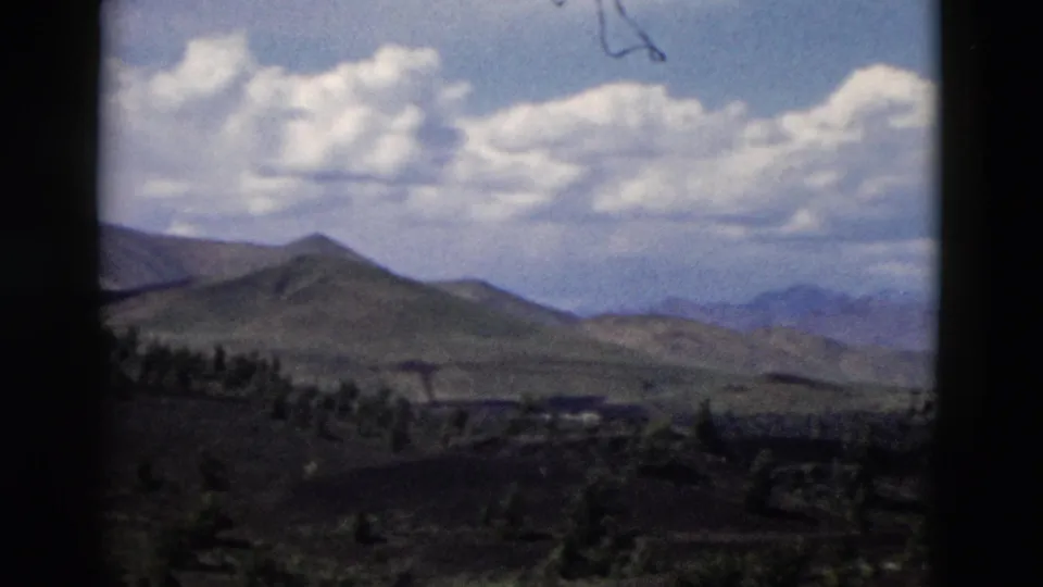 1958 Yellowstone home-movie frame showing rolling hills, distant ridges, dark foreground terrain, and scattered trees under clouds.