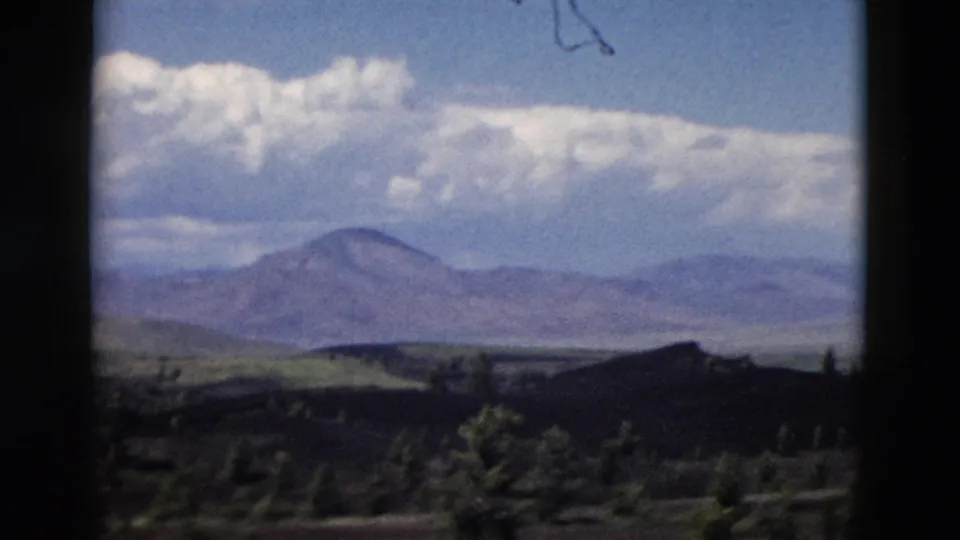 1958 Yellowstone home-movie frame with a rounded distant mountain, layered hills, dark foreground, and bright cloud cover.