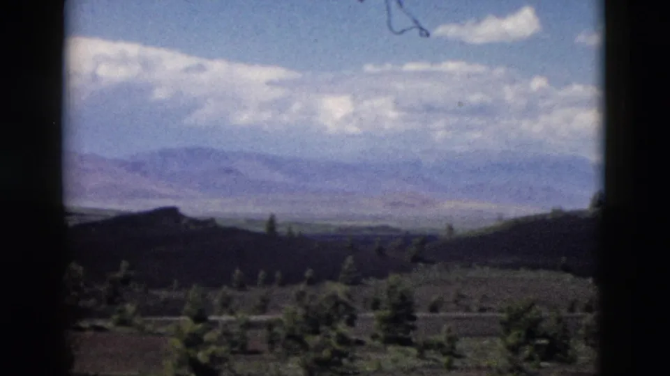 1958 Yellowstone-area home-movie frame showing hazy mountains, layered clouds, dark hills, and foreground evergreens.