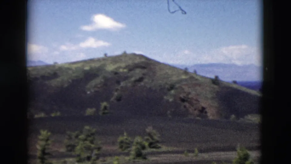 Yellowstone-area 8mm frame of a rounded hill, scattered evergreens, distant mountains, and small clouds.