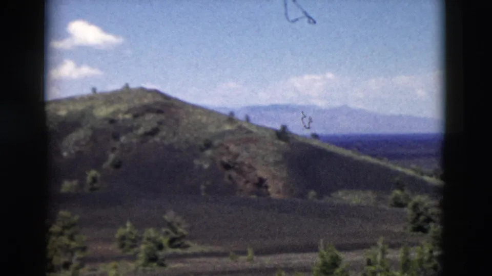 1958 8mm Yellowstone view of a brown hillside, scattered green trees, distant mountains, and blue sky.