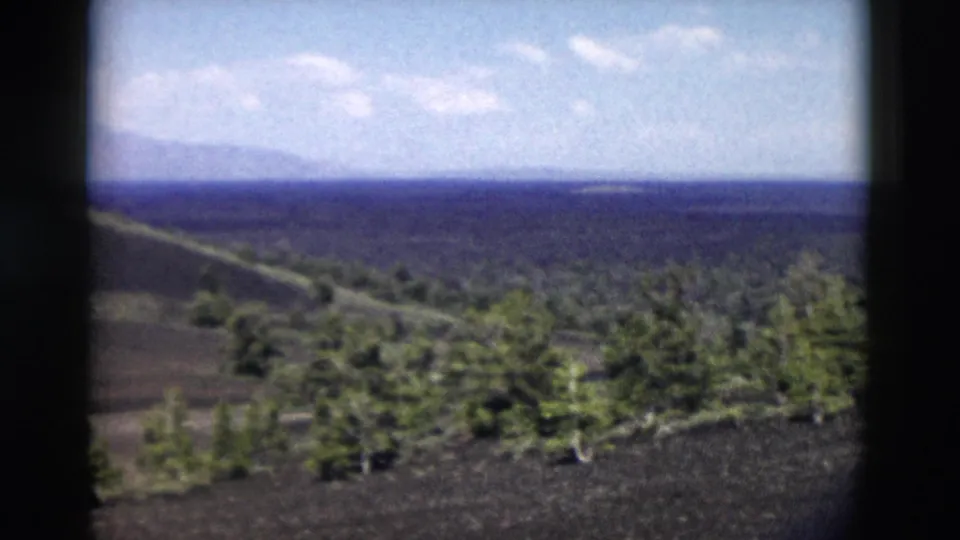 1958 Yellowstone home-movie frame with dark terrain, evergreen trees, and a distant ridge line under clouds.