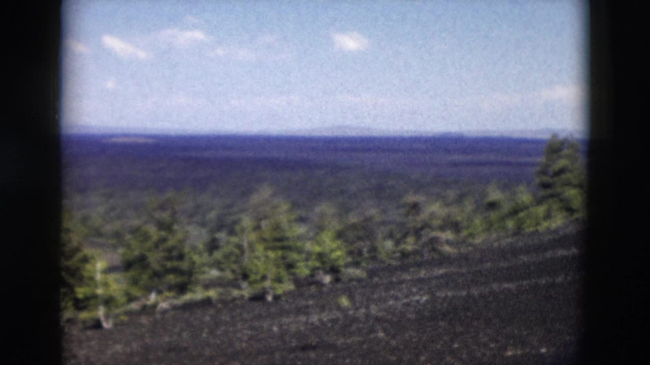 1958 Yellowstone 8mm frame showing a dark volcanic hillside, sparse pines, and a distant forested horizon.