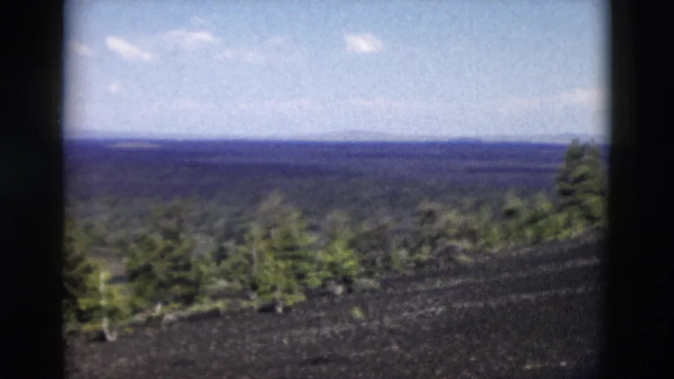 1958 Yellowstone 8mm frame showing a dark volcanic hillside, sparse pines, and a distant forested horizon.