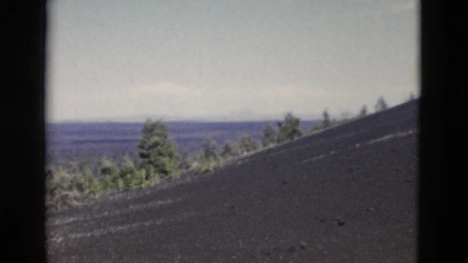1958 Yellowstone home-movie frame with black volcanic ground rising across the foreground and pines on the distant horizon.