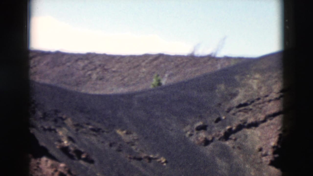 1958 Yellowstone 8mm frame with dark rocky terrain, a small green shrub, and bright sky beyond the ridge.