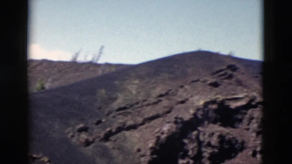 1958 Yellowstone 8mm frame showing a sloped volcanic hillside, rough foreground rock, and pale blue sky.