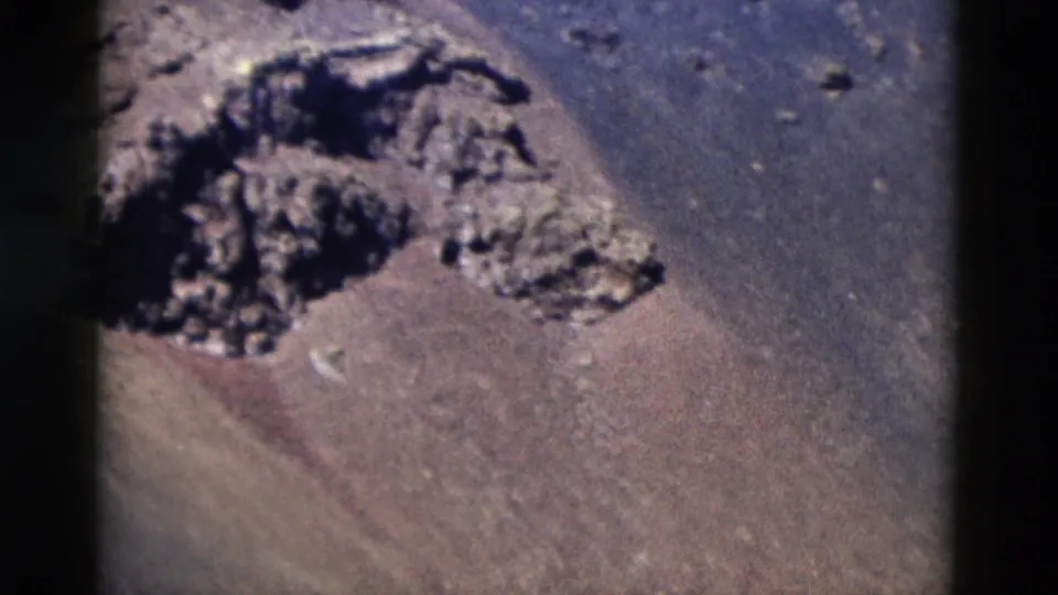 1958 8mm Yellowstone view of a dark, irregular rock outcrop surrounded by dry brown and gray sloping ground.
