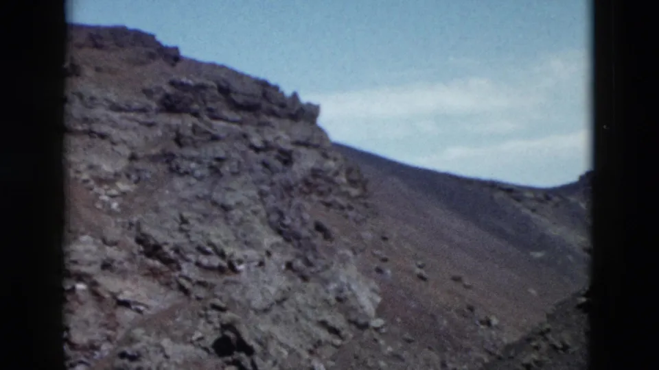 1958 Yellowstone home-movie frame showing a rocky cliff face and barren slope beneath a pale sky, with dark frame edges visible.
