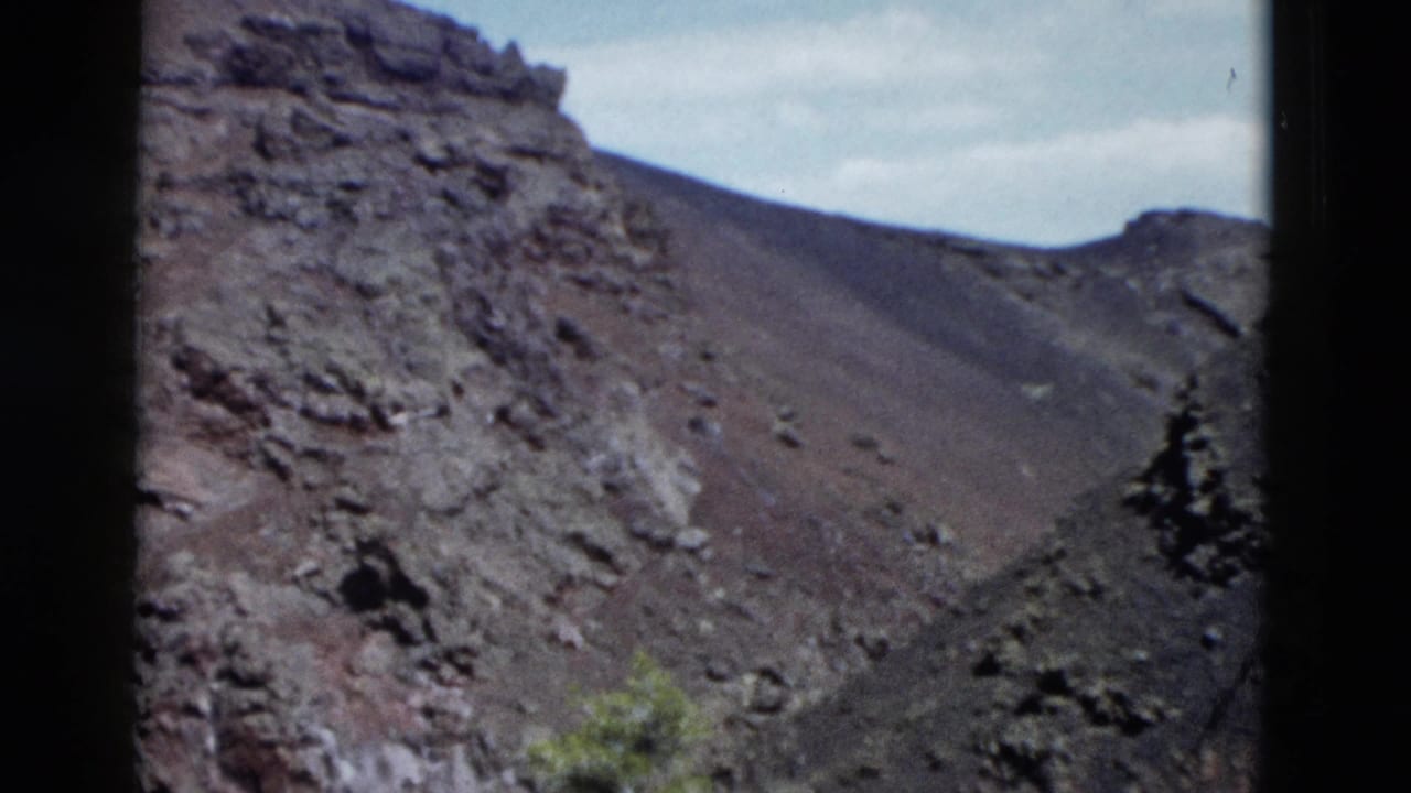 1958 Yellowstone home-movie frame showing a steep rocky slope with dark volcanic-looking soil, scattered stones, sparse vegetation, and film borders.