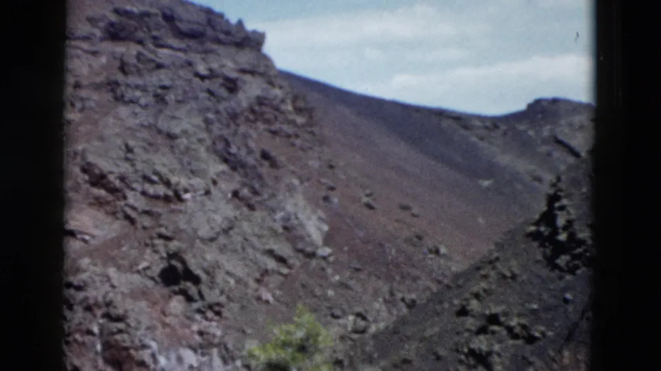 1958 Yellowstone home-movie frame showing a steep rocky slope with dark volcanic-looking soil, scattered stones, sparse vegetation, and film borders.