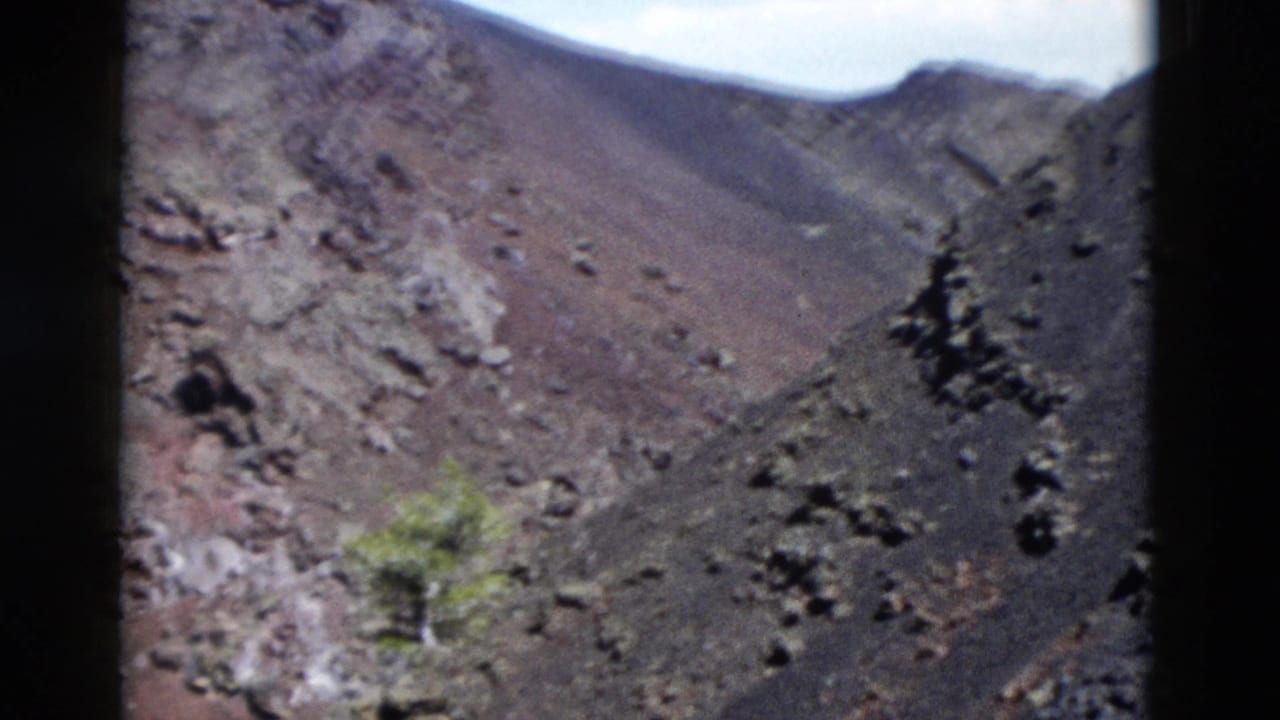 Close view of a barren rocky hillside with reddish soil, loose stones, and a small green shrub.