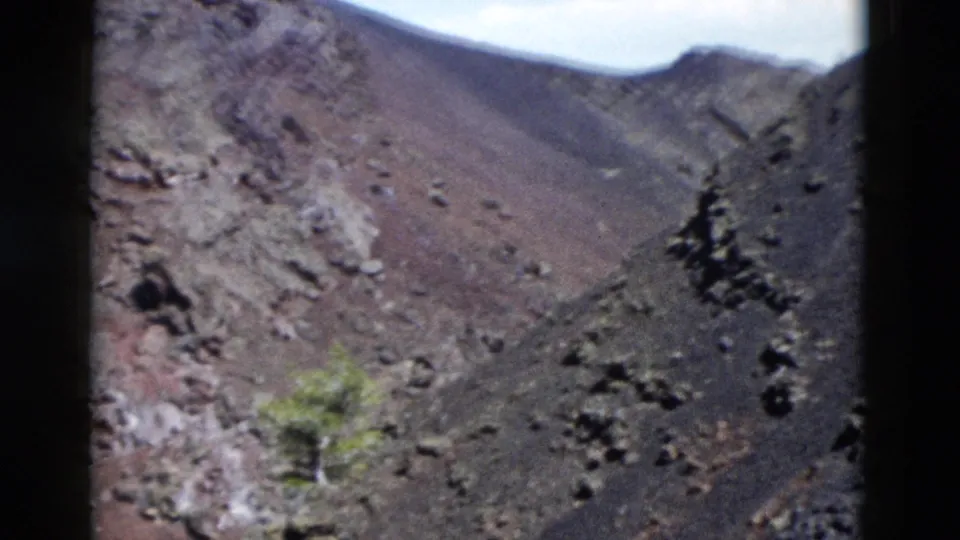 1958 Yellowstone frame with a close handheld view of reddish and dark rocky terrain, loose boulders, a small green shrub, and exposed slope.
