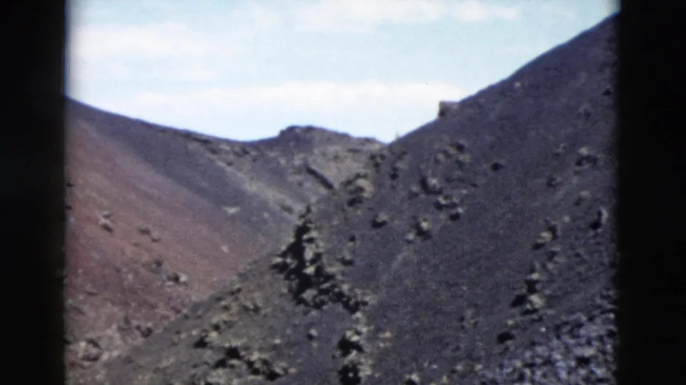 1958 Yellowstone home-movie frame showing dark rocky incline, reddish exposed slope, scattered stones, conifer silhouettes, and pale sky.