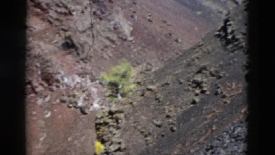 1958 Yellowstone frame showing a shaky close view of rocky mountain terrain, sparse green vegetation, loose stones, and dark film gate borders.