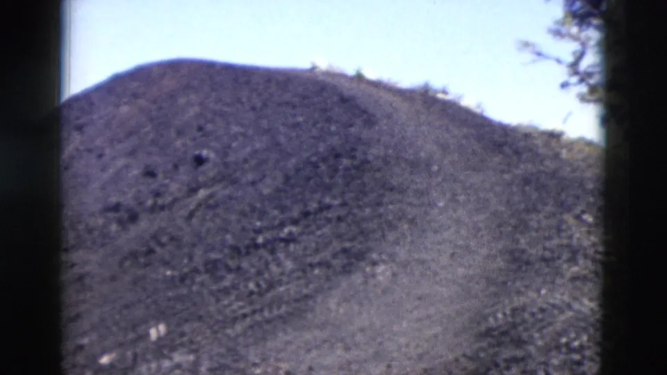 1958 Yellowstone 8mm frame of a dark rocky hillside with sparse plants near the ridge and pale sky in the background.