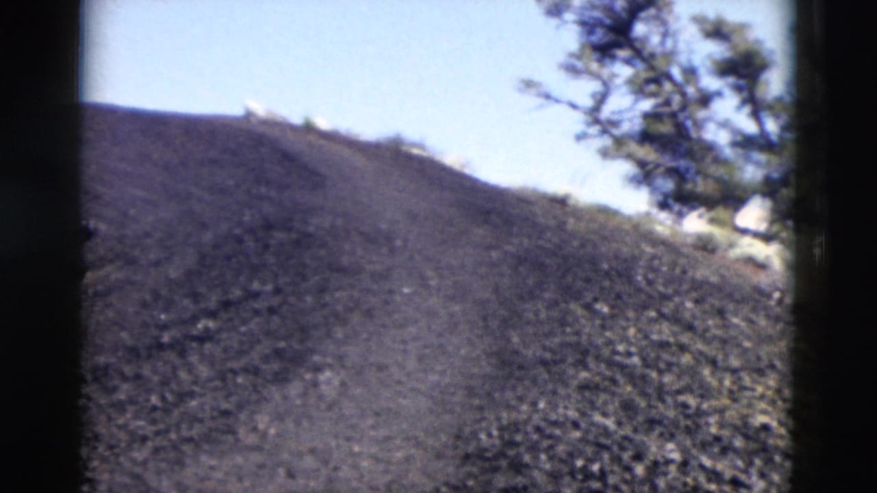 1958 Yellowstone-area 8mm frame showing a dark volcanic-looking slope with sparse pine trees and bright sky.
