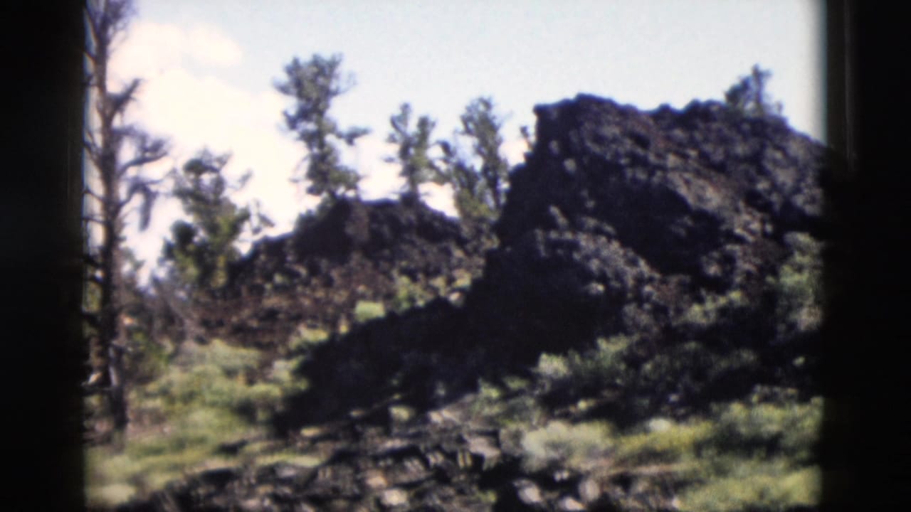 1958 Yellowstone-area 8mm view of dark rock formations, pines, shrubs, and a partly cloudy sky.