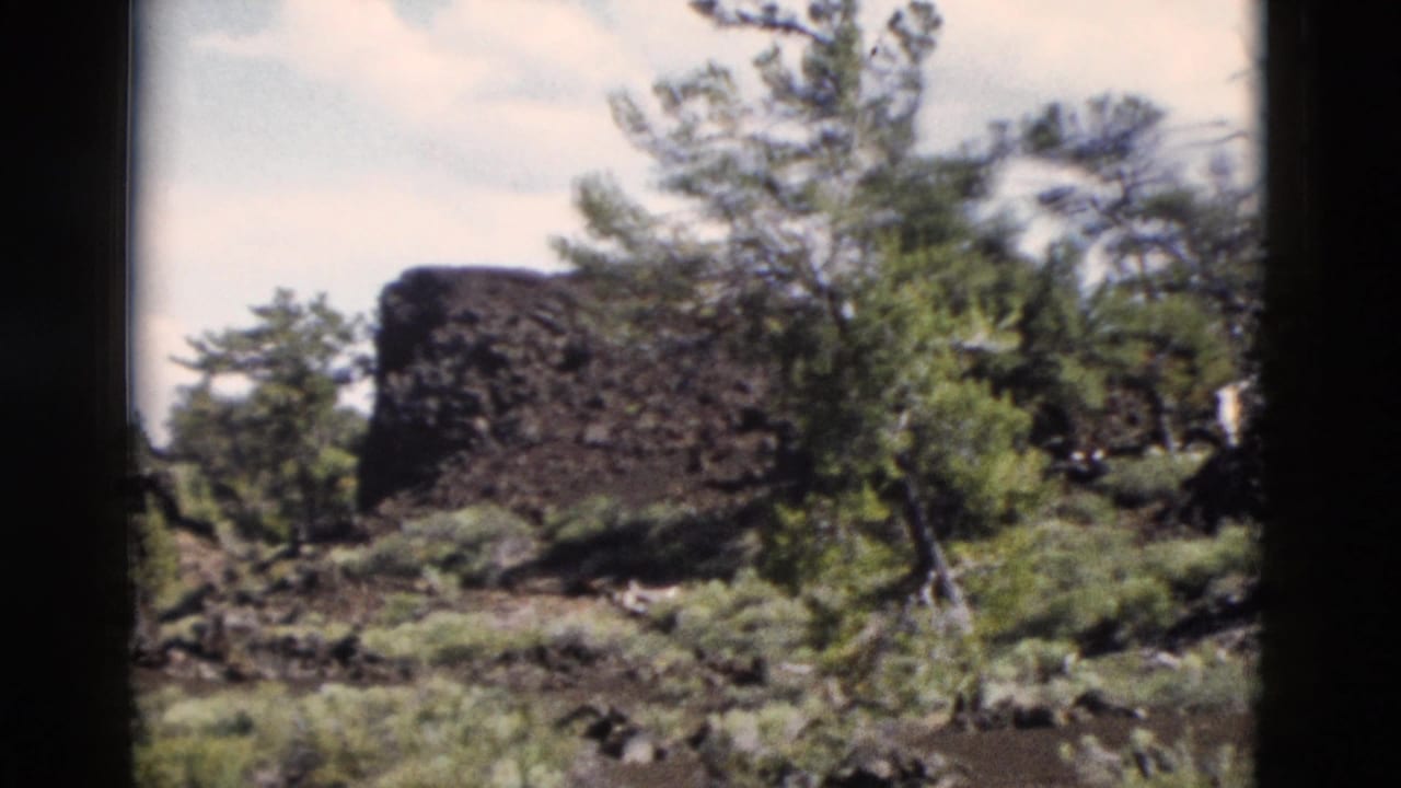 1958 Yellowstone 8mm frame showing a dark rock outcrop amid green shrubs and scattered trees.