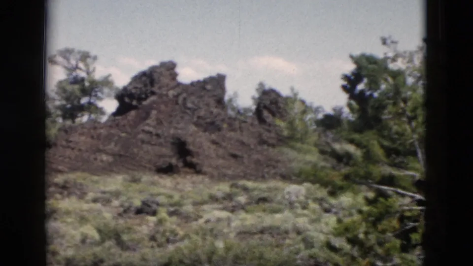1958 Yellowstone home-movie frame of jagged dark rock surrounded by sparse vegetation and trees.