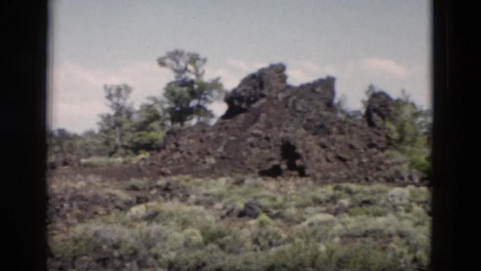 1958 Yellowstone 8mm frame showing dark rock with a small shadowed opening and surrounding shrubs.