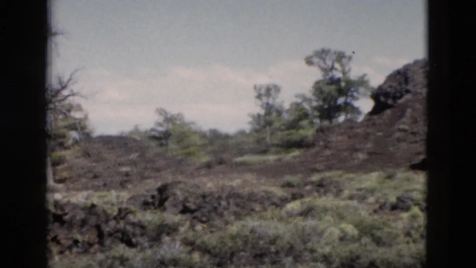 1958 Yellowstone home-movie frame looking across rocky ground, shrubs, trees, and the edge of a dark rock formation.