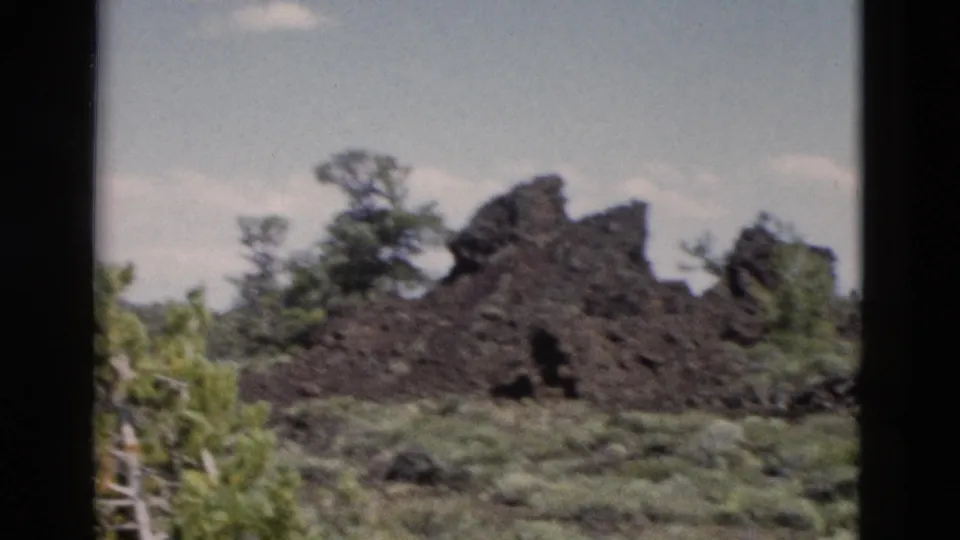 1958 Yellowstone 8mm frame showing a jagged lava outcrop, surrounding grass, trees, and open sky.