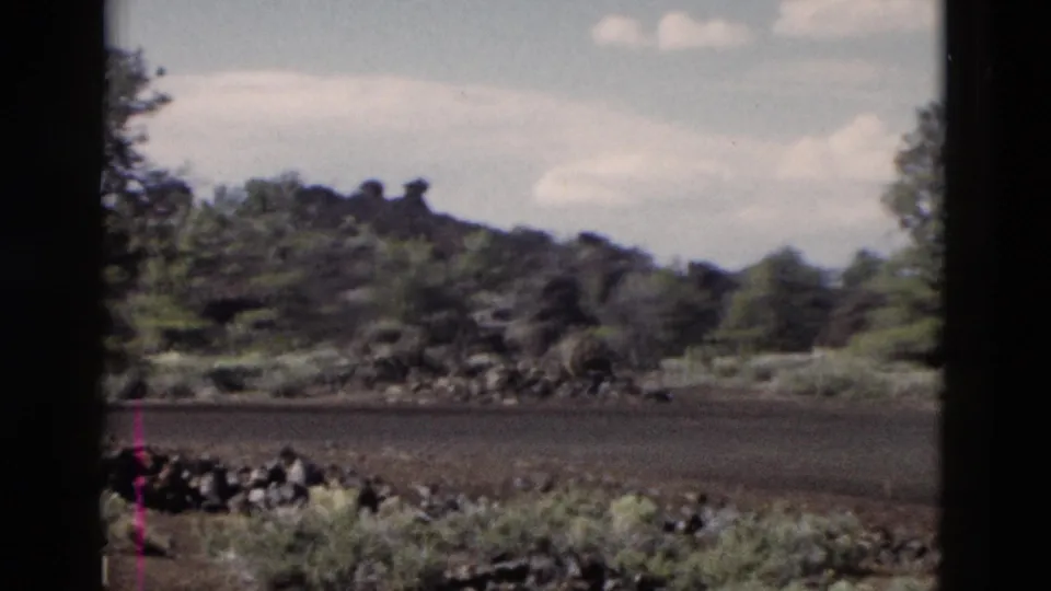 1958 Yellowstone 8mm frame showing dark volcanic terrain, roadside-like foreground, brush, trees, and clouds.