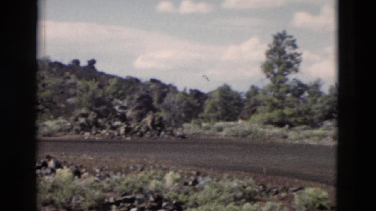 1958 8mm Yellowstone travel view of a paved park road, volcanic-looking rocks, sparse trees, and bright sky framed by dark window edges.