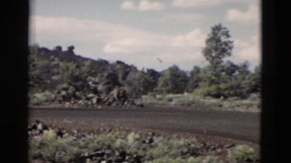 1958 8mm Yellowstone travel view of a paved park road, volcanic-looking rocks, sparse trees, and bright sky framed by dark window edges.