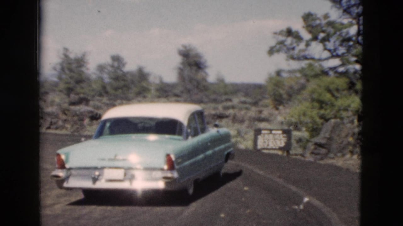 Two-tone classic sedan stopping on a paved overlook road beside rocks and trees.