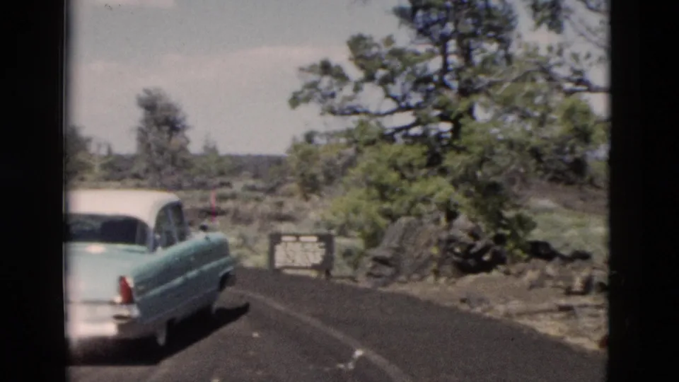 Rear view of a classic two-tone sedan near a Yellowstone roadside sign, with dark pavement, rocks, trees, and low greenery around it.