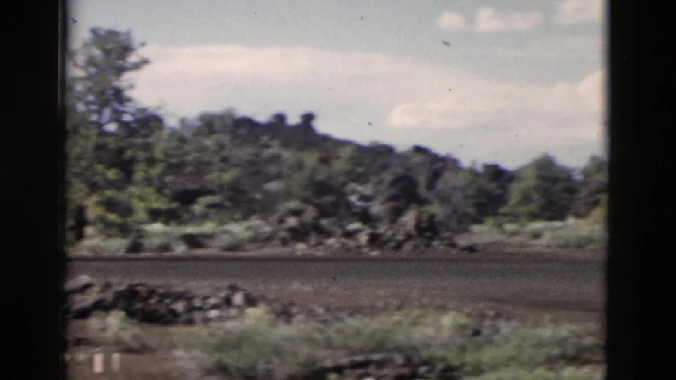End of the Yellowstone pan shows an empty road below dark rocky terrain, scattered shrubs, pines, and soft 8mm grain.