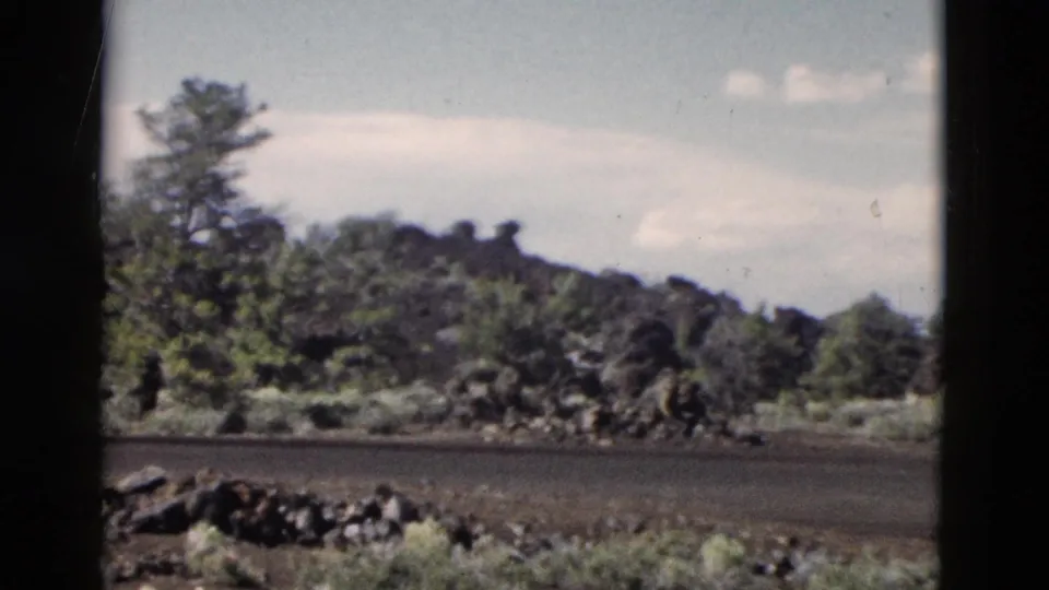 1958 Yellowstone home-movie frame showing a dark secondary road with roadside rocks, shrubs, trees, and a low wooded ridge.