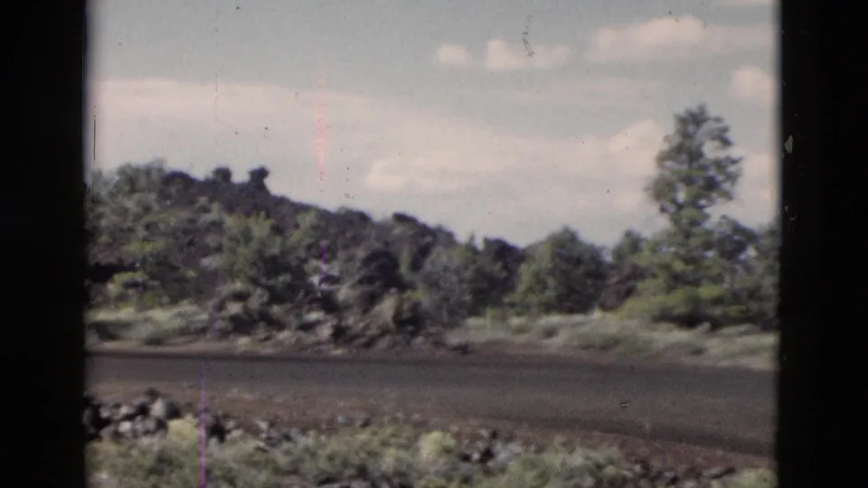 1958 Yellowstone view of a secondary road edge, dark rock formations, low vegetation, and trees in a rural park setting.