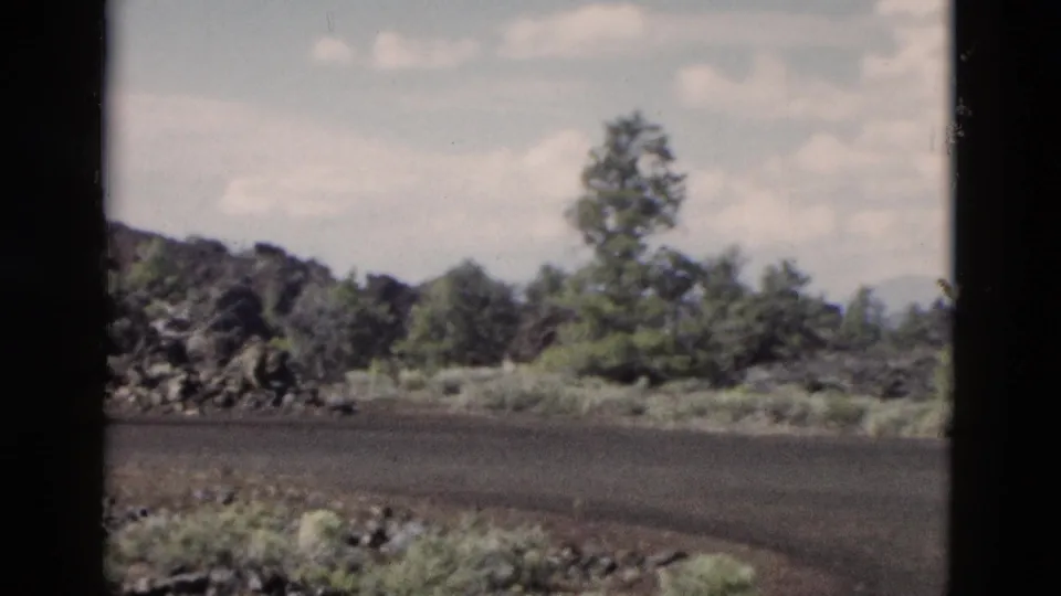 1958 Yellowstone frame from a moving vehicle, showing a dark rural road curving past shrubs, rocks, and trees.