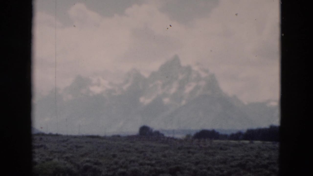 1958 Yellowstone-area home-movie frame with snowy peaks, muted clouds, scrub foreground, and visible 8mm frame edges.