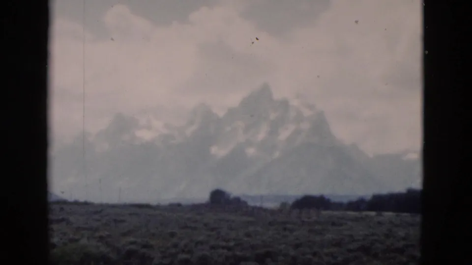 1958 Yellowstone-area home-movie frame with snowy peaks, muted clouds, scrub foreground, and visible 8mm frame edges.