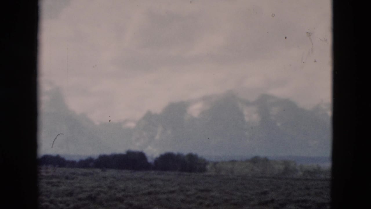 1958 8mm view of the Grand Teton range from the Yellowstone, Wyoming metadata area, framed by dark vertical window edges over a muted plain.
